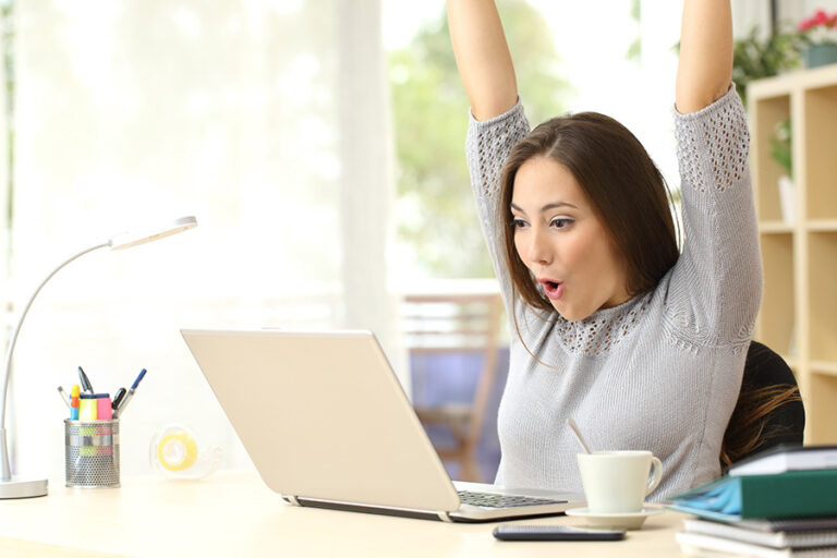 excited woman sitting at her desk looking at laptop