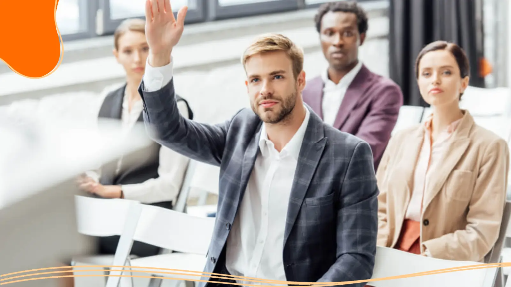 A young office worker, at a meeting about a business card online order, holds his hand up to ask a question.