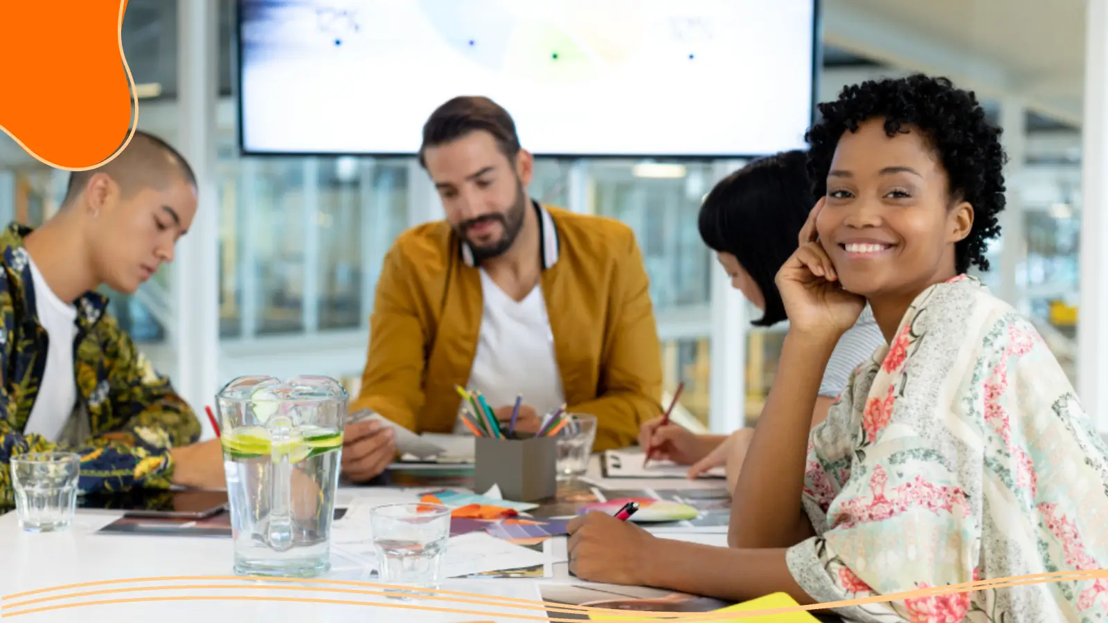 A young woman with short curly hair smiles at the camera, resting her chin on her hand at a modern conference table while colleagues review charts behind her, an example of branding for small businesses in a collaborative workspace.