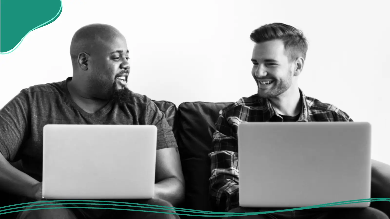 A black-and-white photograph of two men sitting on a sofa side by side, each holding open laptops on their laps and smiling as they turn to look at each other, reflecting collaboration to build your brand.