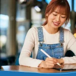 A smiling woman using a stylus on a tablet while sitting at a table in a cafe, representing build your brand through digital creativity.
