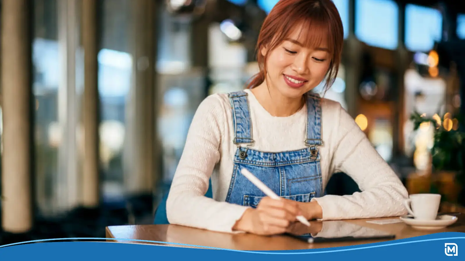A smiling woman using a stylus on a tablet while sitting at a table in a cafe, representing build your brand through digital creativity.