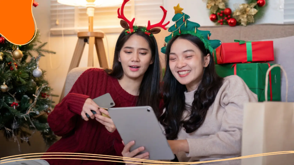 Two happy friends wearing festive headbands look at a tablet together to send digital holiday greetings to best friends, surrounded by Christmas gifts.