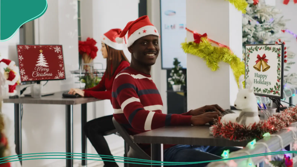 Smiling man wearing a Santa hat typing at his desk to send digital holiday greetings for best friends from a decorated office setting.