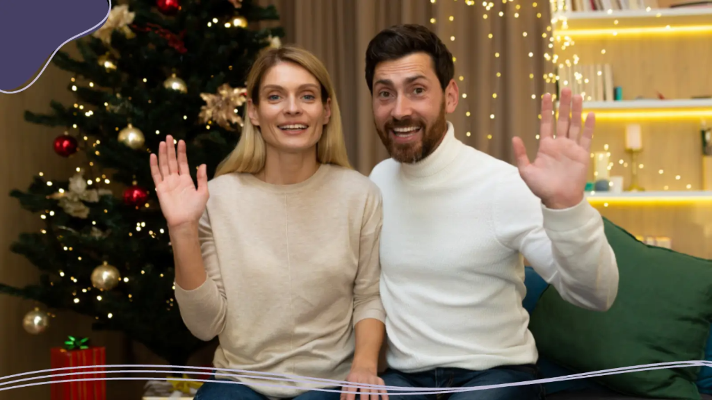 Smiling couple sitting by an illuminated Christmas tree and waving at the camera while sharing virtual holiday greetings for best friends on a video call.