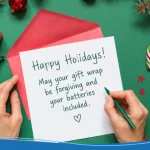 Handwriting funny holiday card messages on a white card with red envelope, featuring the text "Happy Holidays! May your gift wrap be forgiving and your batteries included," surrounded by gifts and candy canes.