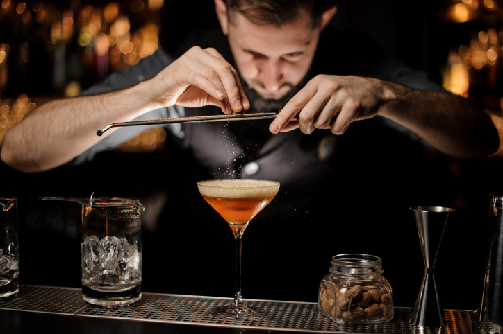 Professional bartender adding to a cocktail in the glass with a whipped cream a grated nutmeg